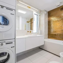 Nowy Wymiar Mebli - Nowoczesna łazienka z pralką i suszarką w zabudowie, białe meble łazienkowe z umywalką nablatową, złote płytki heksagonalne i armatura.