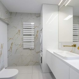 Nowy Wymiar Mebli - Nowoczesna łazienka w Niepołomicach z białymi płytkami imitującymi marmur, złotą armaturą, podwieszaną toaletą i pralką w zabudowie.