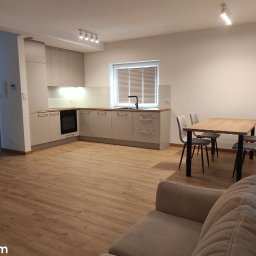 Nowy Wymiar Mebli - Jasna kuchnia w narożniku z drewnianym blatem, widok na stół z czterema krzesłami i fragment sofy w salonie, oświetlenie sufitowe.