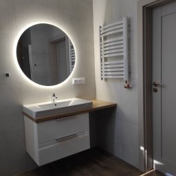 Nowy Wymiar Mebli - Nowoczesna łazienka z okrągłym lustrem podświetlanym LED, białą szafką pod umywalką z dwiema szufladami i grzejnikiem drabinkowym na szarej płytce.