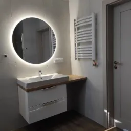 Nowoczesna łazienka z okrągłym lustrem podświetlanym LED, białą szafką pod umywalką z dwiema szufladami i grzejnikiem drabinkowym na szarej płytce.