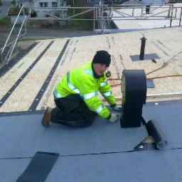 Dekarz w neonowej kurtce montuje czarny kominek wentylacyjny na dachu pokrytym papą. Widoczna podbudowa dachu z płyt OSB i membrana dachowa.