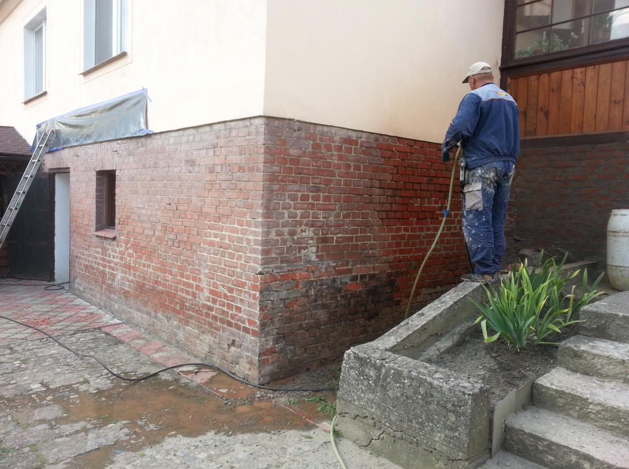 Czyszczenie ceglanej elewacji budynku z wykorzystaniem myjki ciśnieniowej. Widoczny murarz w roboczym ubraniu i fragment elewacji po czyszczeniu.