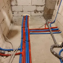 Nowa instalacja wodna z rurami w kolorze niebieskim i czerwonym, zabezpieczonymi pianką montażową, w surowym wnętrzu budynku z bloczków.