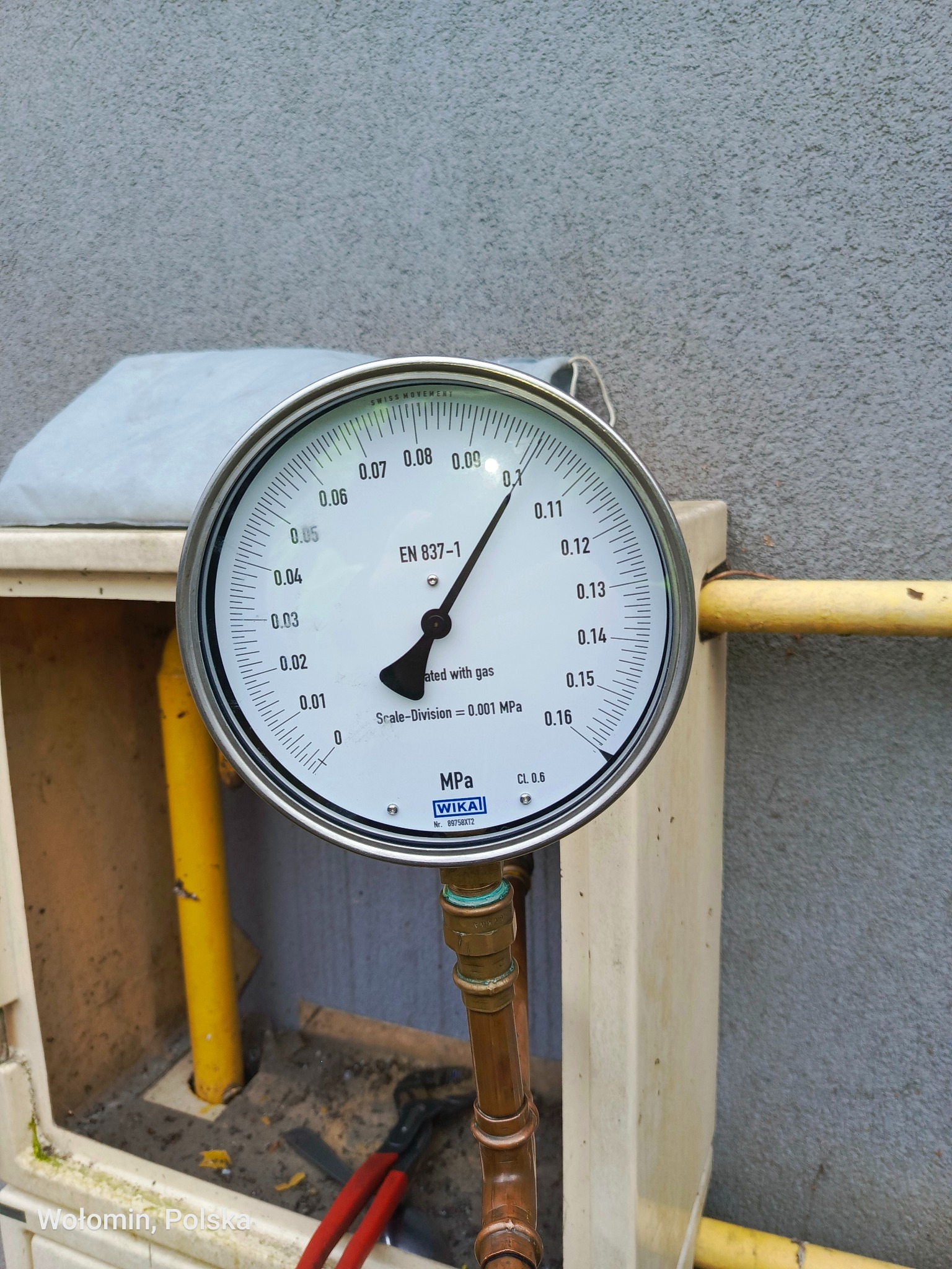 Manometr WIKA na instalacji gazowej w skrzynce, wskazujący ciśnienie. Widoczne miedziane rury i żółty gazociąg. W tle szara ściana.