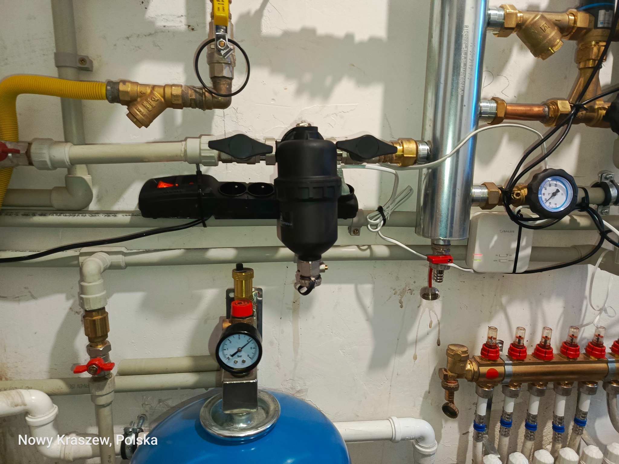 Złożona instalacja grzewcza z widocznym rozdzielaczem, filtrem, manometrami i zaworami. Żółty przewód gazowy, białe rury PCV i miedziane elementy. Całość na tle ściany.