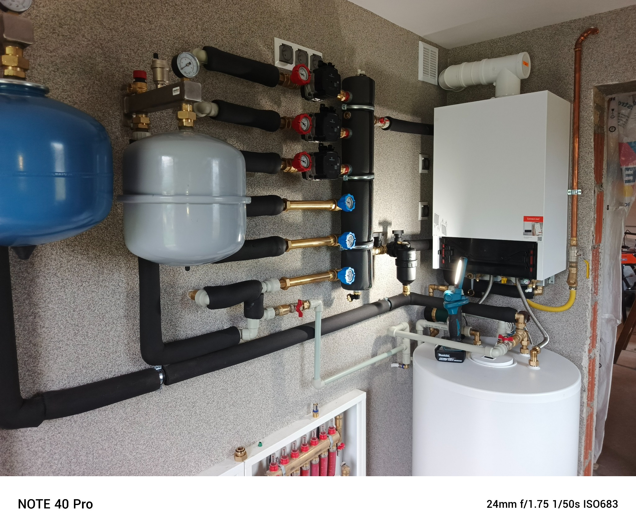 Złożona instalacja grzewcza: biały piec gazowy, zbiorniki wyrównawcze (niebieski i szary), czarne rury izolowane oraz mosiężne zawory. Elementy instalacji podłogowej widoczne w dolnej części.