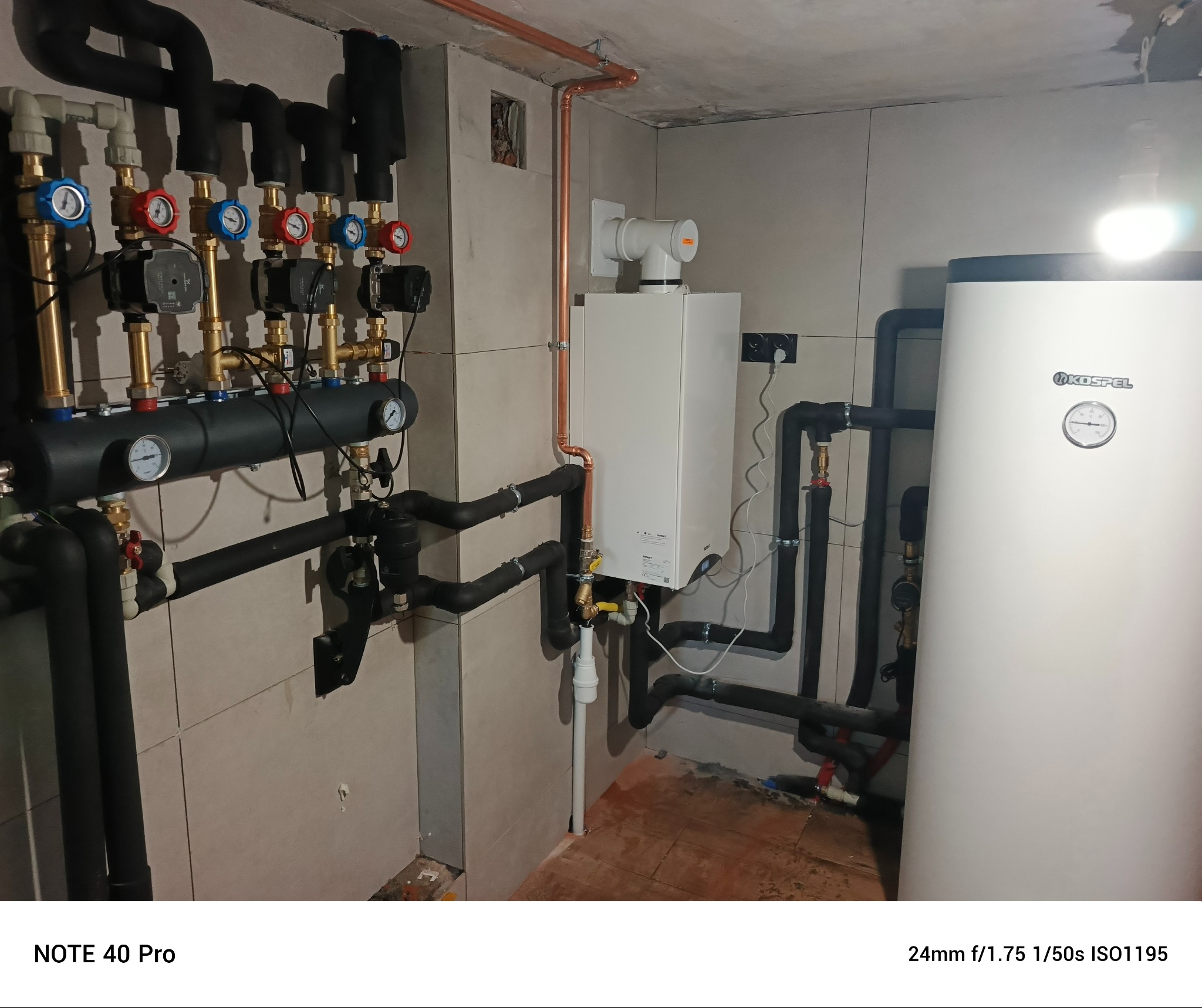 Kotłownia z piecem gazowym, zasobnikiem ciepłej wody i rozdzielaczem. Miedziane rury, czarne rury instalacyjne. Białe płytki na ścianach.