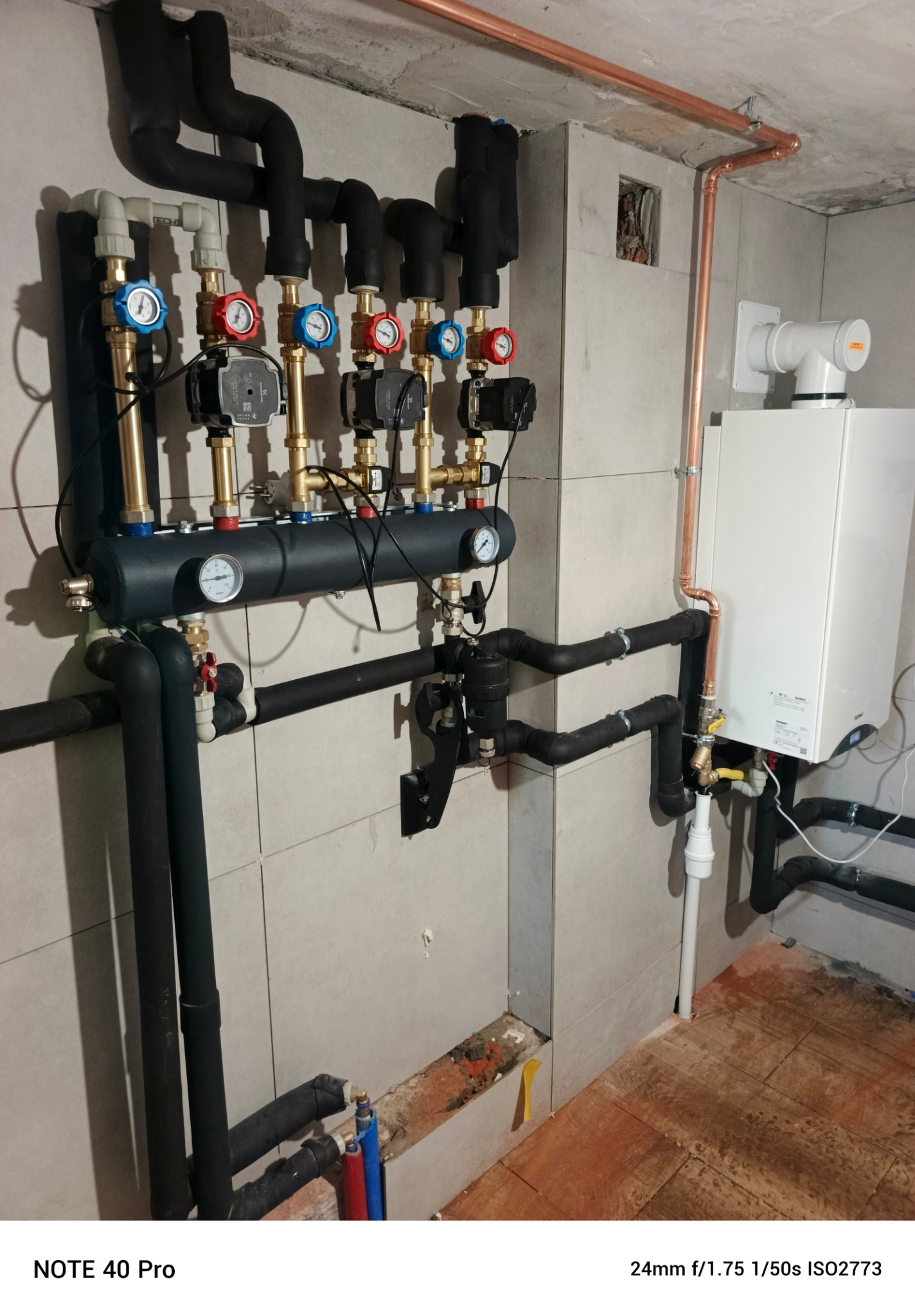 Kompleksowa instalacja grzewcza z kotłem gazowym, rozdzielaczem i rurami miedzianymi na tle jasnych płytek. Widoczne manometry i pompy obiegowe. Detal instalacji.