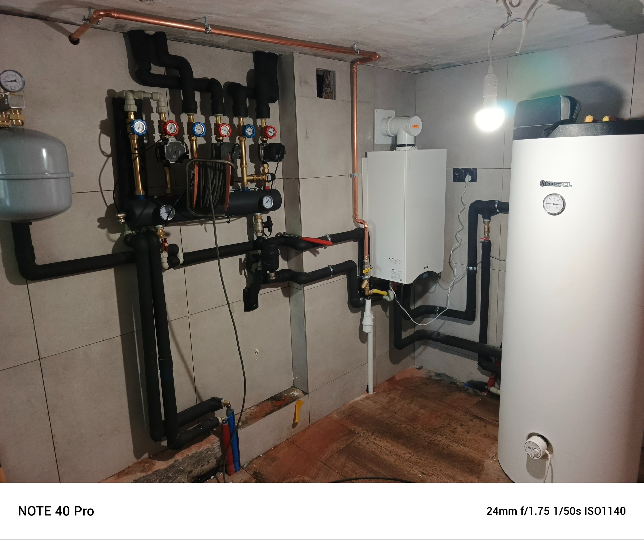 Instalacja centralnego ogrzewania z kotłem gazowym, zbiornikiem i rozdzielaczem. Widoczne rury miedziane i czarne izolowane. Całość zamontowana na tle szarych płytek.