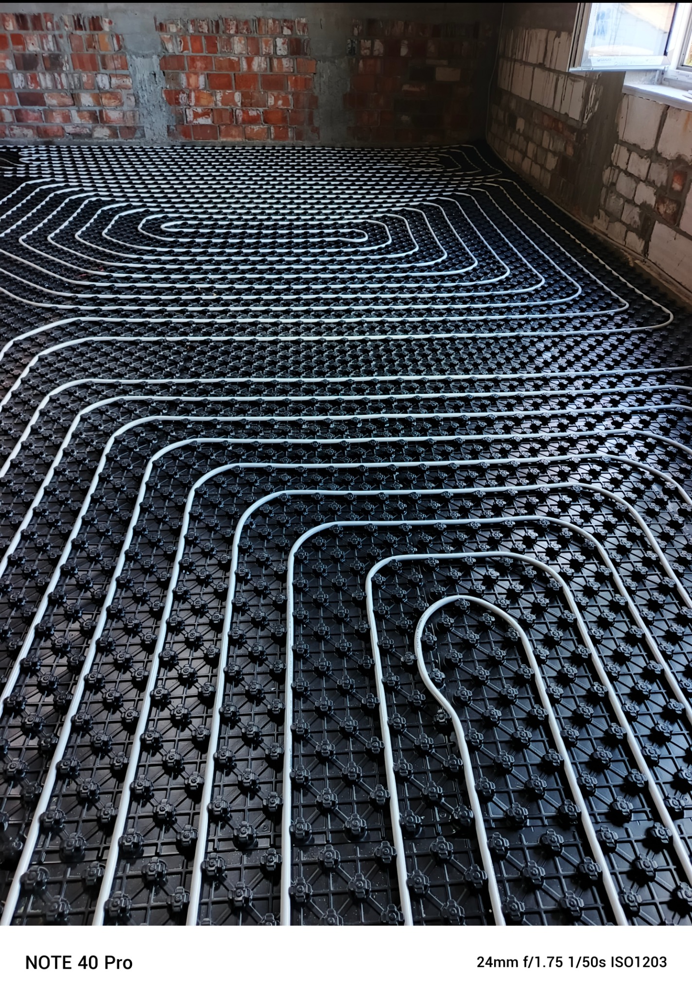 Białe rury ogrzewania podłogowego ułożone na czarnej macie systemowej, widok z góry, surowa ściana z cegły w tle, pokój w trakcie wykończenia.