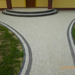 Jasna kostka brukowa tworząca ścieżkę rozwidlającą się od schodów wejściowych, obrzeża z ciemnej kostki, trawnik po bokach.