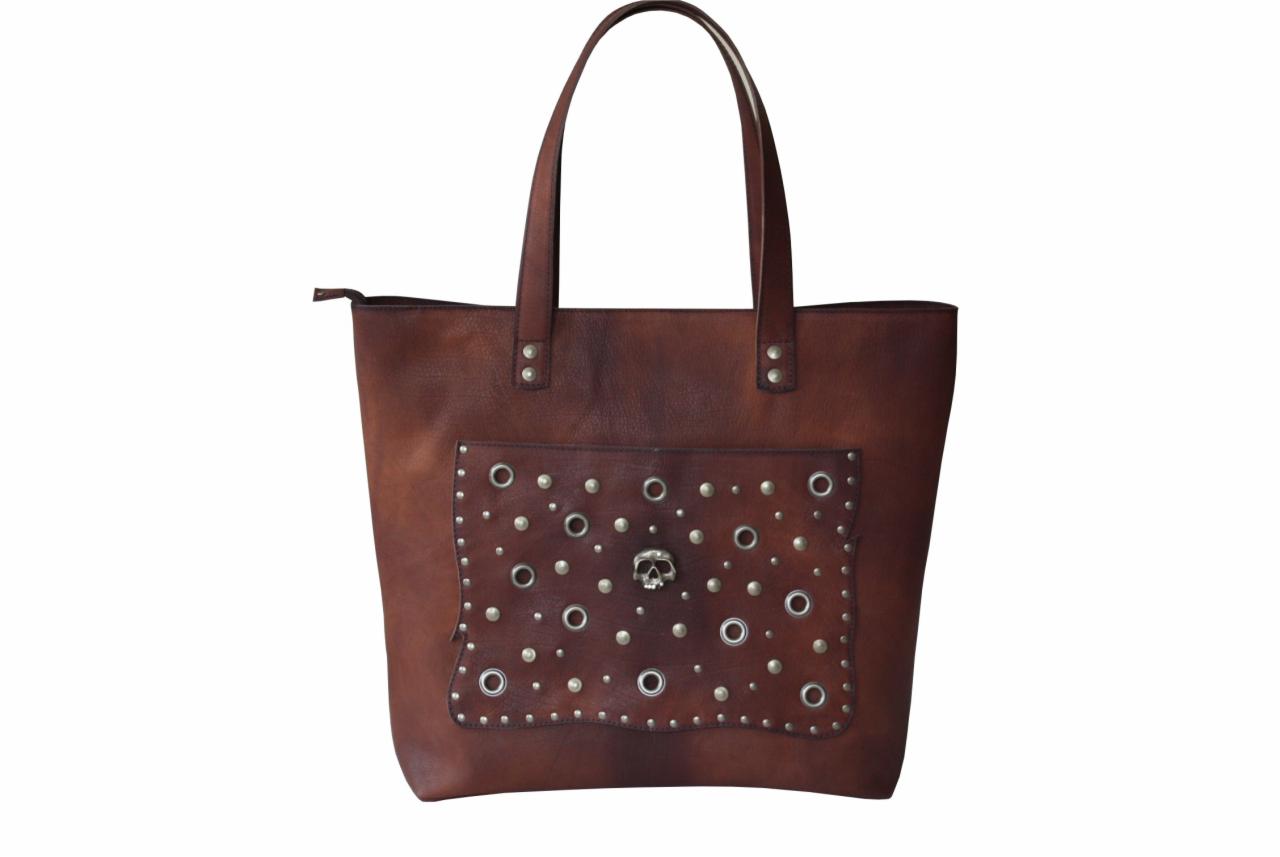 Brązowa torba typu shopper ze skóry z kieszenią zdobioną srebrnymi nitami i czaszką pośrodku, na białym tle.