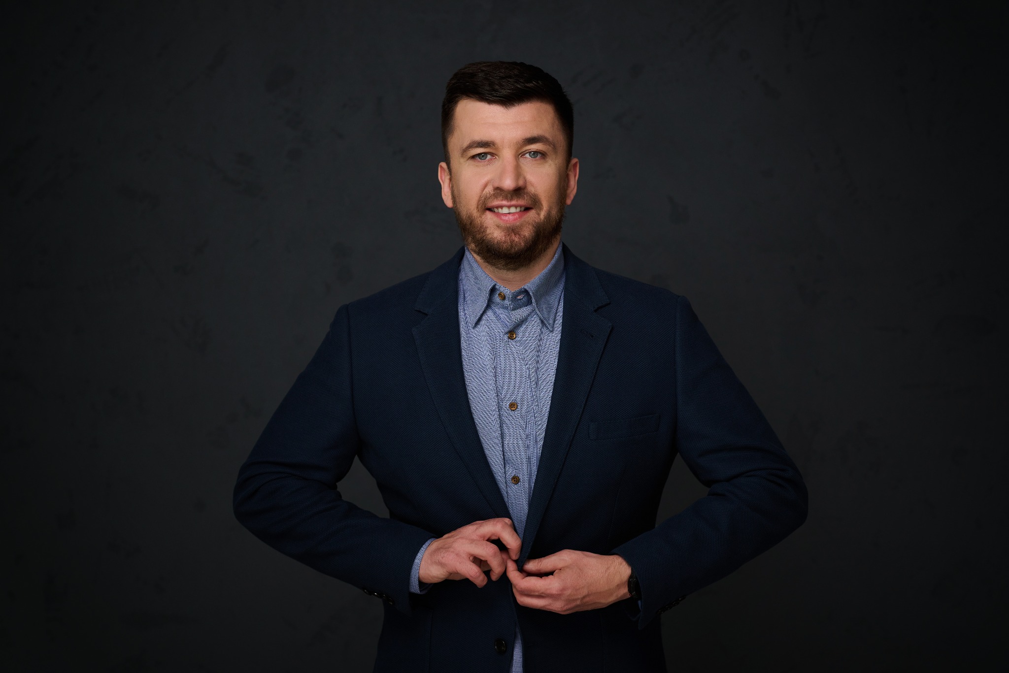 Mężczyzna z brodą, w niebieskiej koszuli i marynarce, zapina guzik. Portret na ciemnoszarym tle, z delikatnym uśmiechem i spojrzeniem w obiektyw.