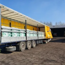 Ciężarówka z otwartą naczepą załadowana workami z materiałem opałowym, widoczna na tle magazynu i placu składowego w słoneczny dzień.