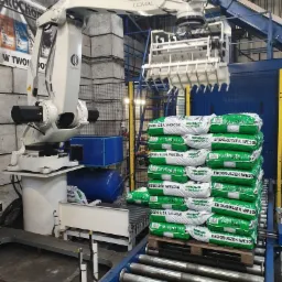 Zautomatyzowany proces paletyzacji ekogroszku: robot przemysłowy Comau układa worki na palecie na linii produkcyjnej.