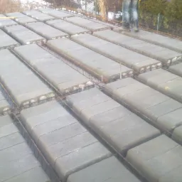 Strop gęstożebrowy w trakcie budowy, widoczne betonowe belki i zbrojenie stalowe, na górze stoją robotnicy w jeansach i roboczych butach.