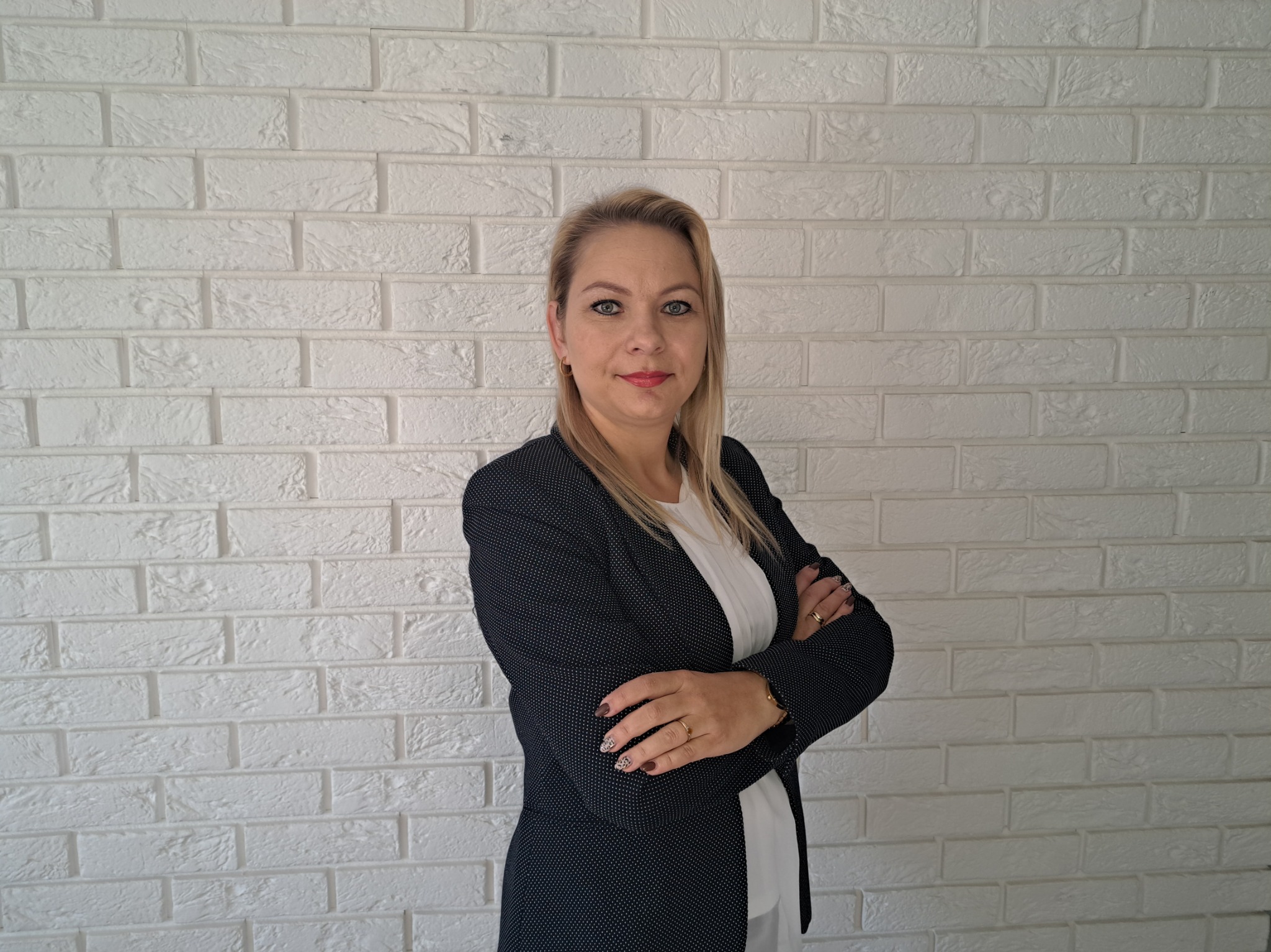 Portret kobiety w czarnej marynarce na tle białej ściany z cegieł, z założonymi rękami i delikatnym uśmiechem.