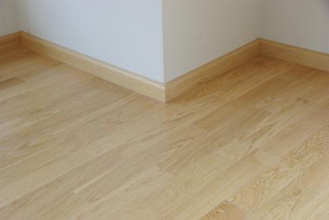 Jasny parkiet ułożony w rogu pomieszczenia, z widocznymi listwami przypodłogowymi w kolorze drewna.