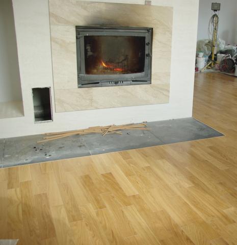 Ułożony parkiet w odcieniach jasnego drewna, widoczny kominek z paleniskiem i przygotowane listewki przypodłogowe.