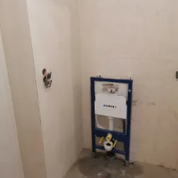 Stelaż podtynkowy Geberit zamontowany w surowym wnętrzu łazienki, widoczne instalacje elektryczne i wodne, ściany przygotowane do dalszych prac wykończeniowych.