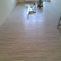 Jasna podłoga z paneli ułożona wzdłuż dłuższego boku pomieszczenia, widoczne niedbale odłożone narzędzia i odkurzacz, ściany przygotowane do malowania.