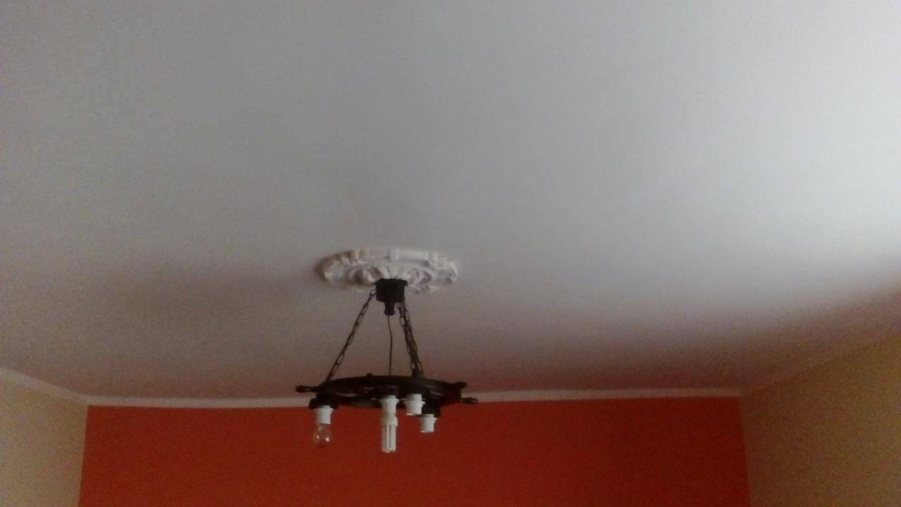 Biały sufit z dekoracyjną rozetą i wiszącą, stylizowaną lampą z żarówkami, pomarańczowa ściana w tle.