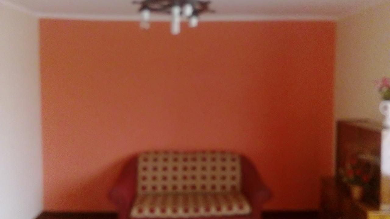 Ściana w odcieniach pomarańczy po malowaniu, widoczny fragment sofy i lampy sufitowej, słabe oświetlenie.