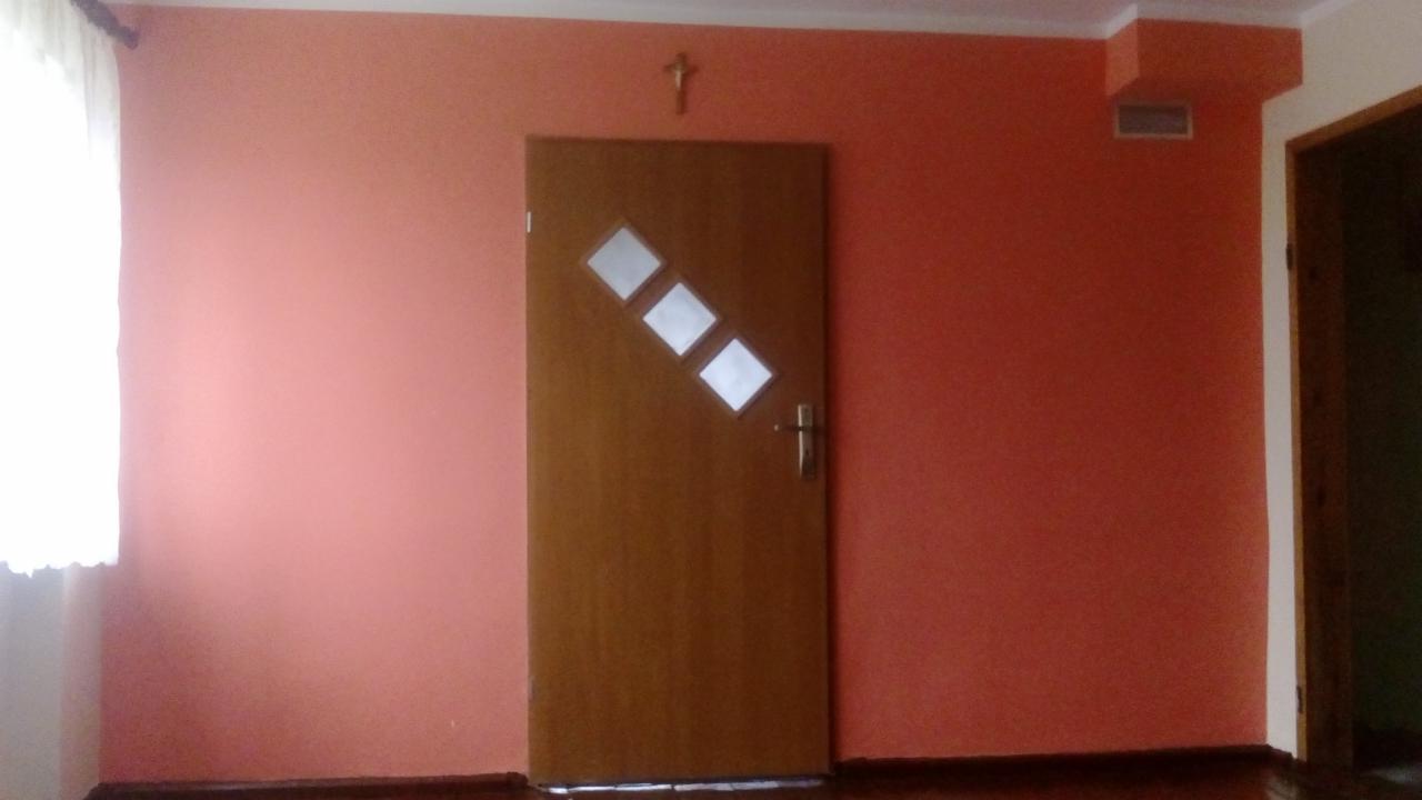 Ściana pomalowana na kolor brzoskwiniowy, drewniane drzwi z trzema romboidalnymi okienkami, krzyż na ścianie, widoczny fragment podłogi i okna.