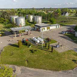 MAC-BENZ MACIEJ I BARBARA KŁACZKIEWICZ SPÓŁKA JAWNA - Widok z góry na stację paliw z dystrybutorami, zbiornikami magazynowymi i budynkiem obsługi, otoczoną zielenią i zabudowaniami mieszkalnymi w tle.