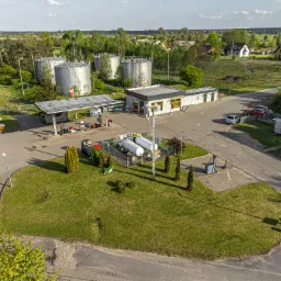 Widok z góry na stację paliw z dystrybutorami, zbiornikami magazynowymi i budynkiem obsługi, otoczoną zielenią i zabudowaniami mieszkalnymi w tle.
