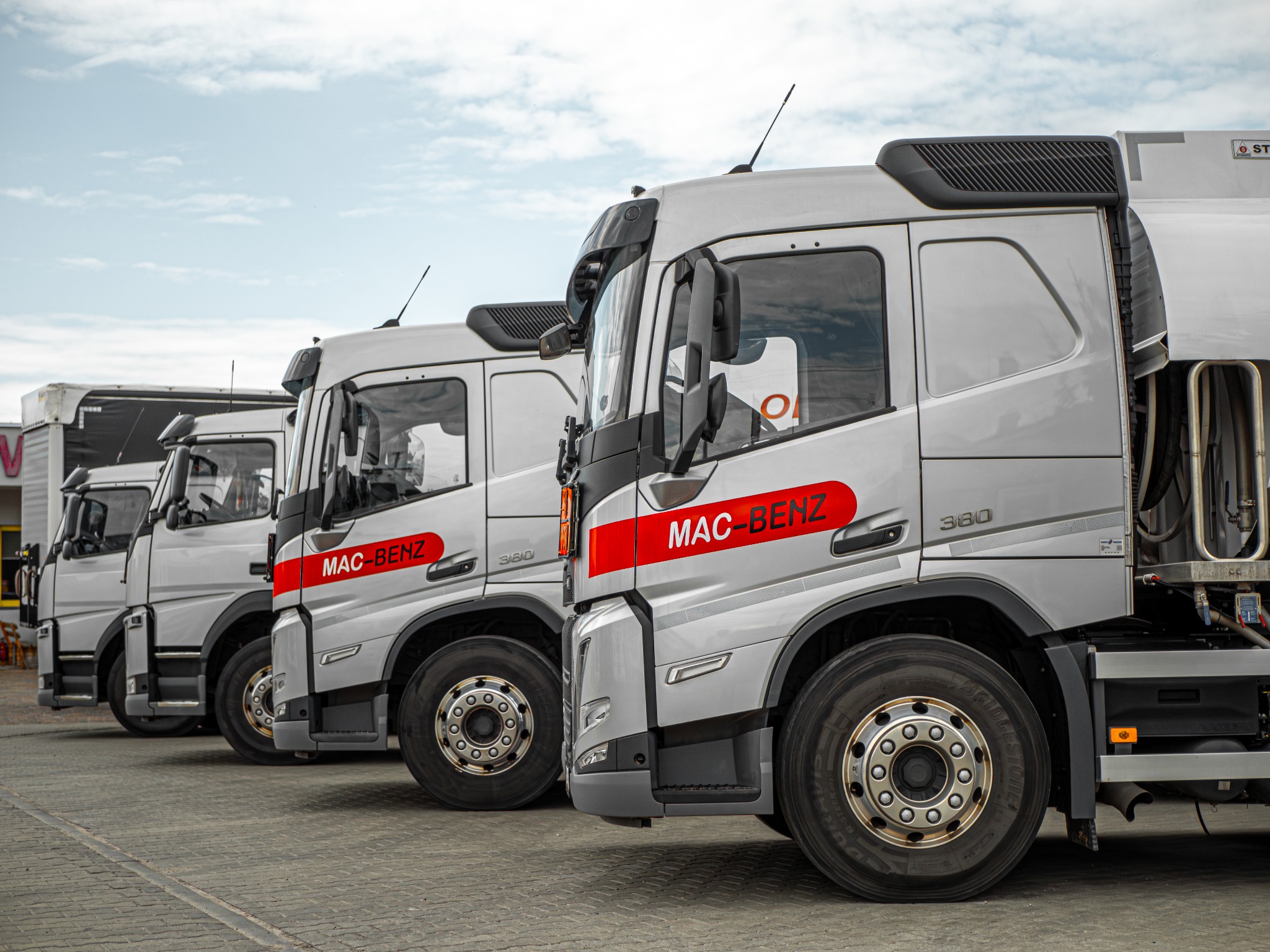 Rząd cystern transportowych z logo MAC-BENZ 380, zaparkowanych na betonowym placu pod pochmurnym niebem.