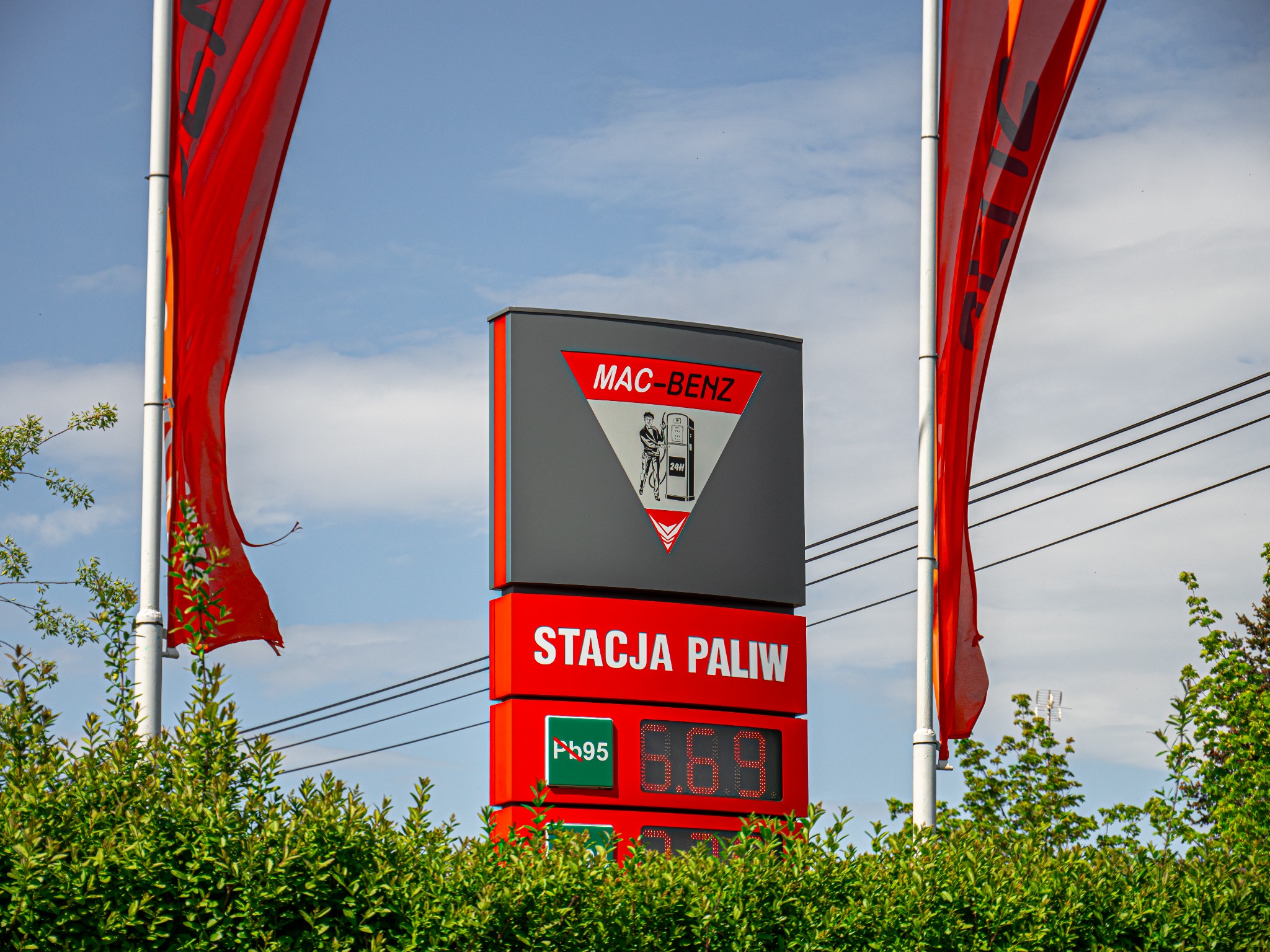 Pylon reklamowy stacji paliw Mac-Benz z aktualnymi cenami paliw, widoczny zza zielonych krzewów i czerwonych flag na tle pochmurnego nieba.