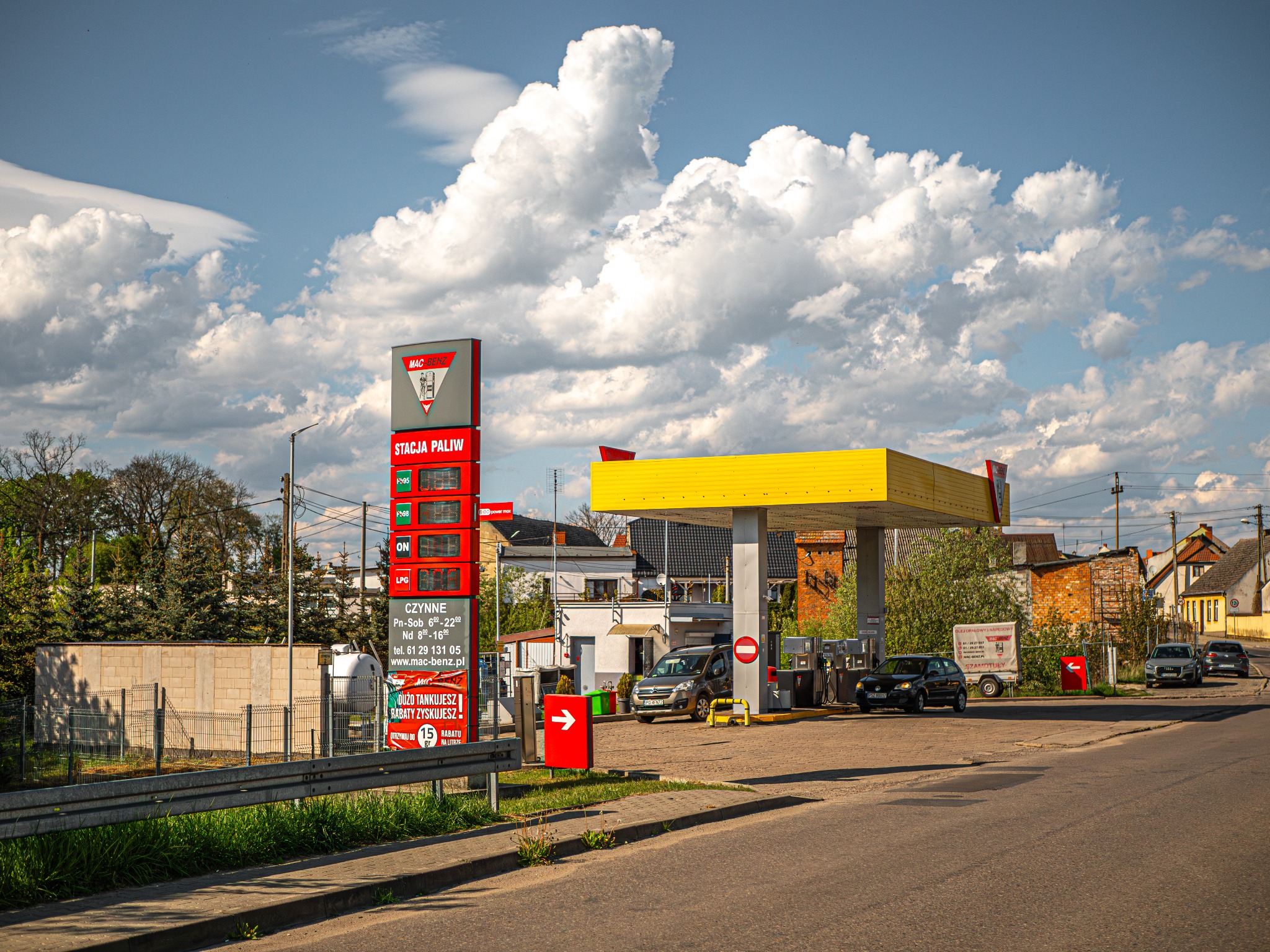 Stacja paliw z żółtym zadaszeniem dystrybutorów, widoczny pylon z cenami paliw i logo, kilka samochodów na podjeździe, błękitne niebo z białymi chmurami w tle.