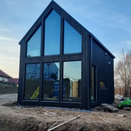 Szymwood - Nowoczesny, czarny dom z dużymi oknami, w trakcie budowy, widoczne materiały budowlane i niezagospodarowany teren wokół.