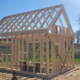 Wznoszenie drewnianej konstrukcji szkieletowej domu na fundamencie, widoczne narzędzia budowlane i otaczający krajobraz.