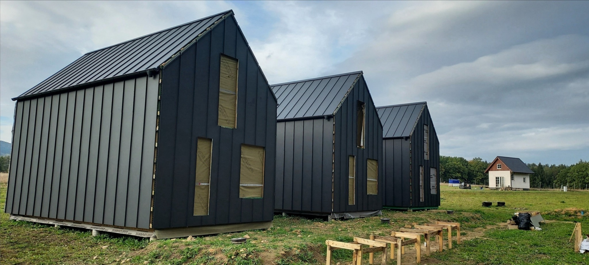 Trzy czarne domy w stylu nowoczesnej stodoły, z widocznymi otworami okiennymi zabezpieczonymi folią, ustawione w rzędzie na trawiastym terenie, z widocznymi elementami konstrukcji pod taras i małym...