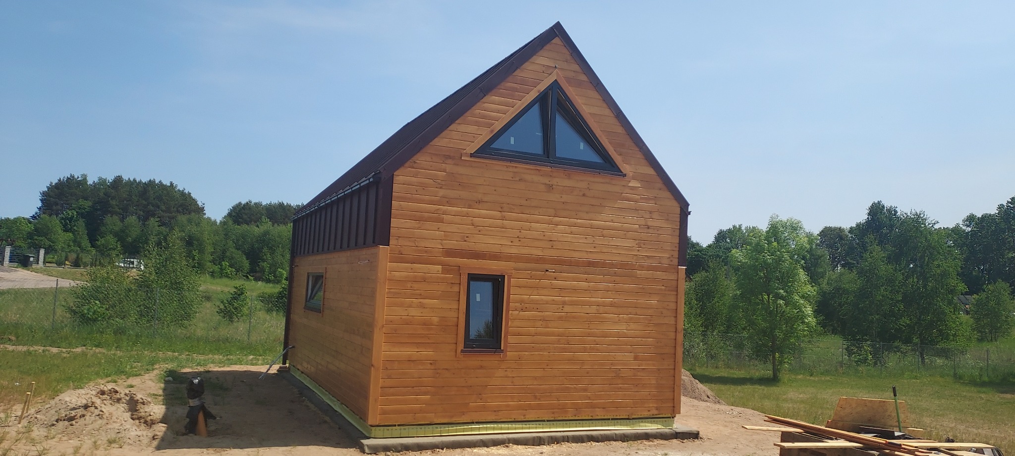 Nowoczesny, parterowy dom z drewnianą elewacją w trakcie budowy, z charakterystycznym trójkątnym oknem na szczycie, widoczne elementy konstrukcyjne i przygotowany teren.