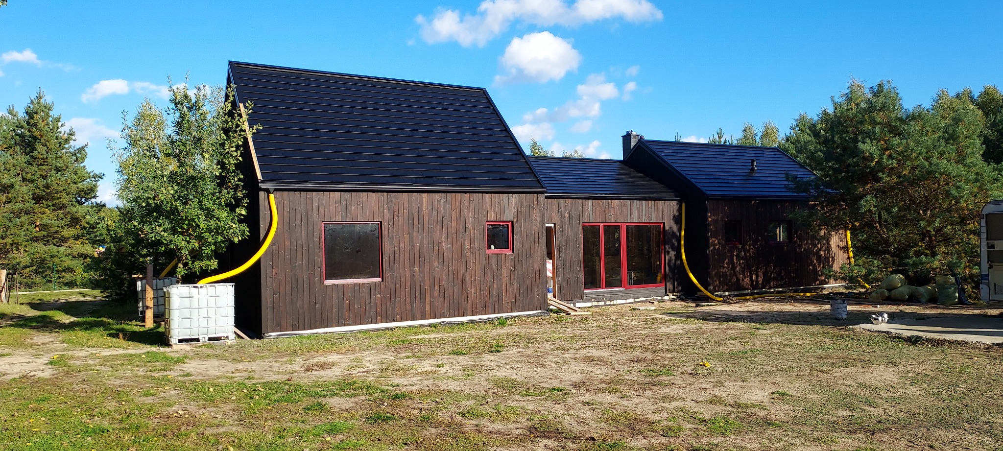 Nowoczesny, parterowy dom z ciemnego drewna z czarnym dachem, czerwonymi ramami okiennymi i żółtą rurą odprowadzającą wodę deszczową do zbiorników na wodę, otoczony drzewami i trawnikiem.