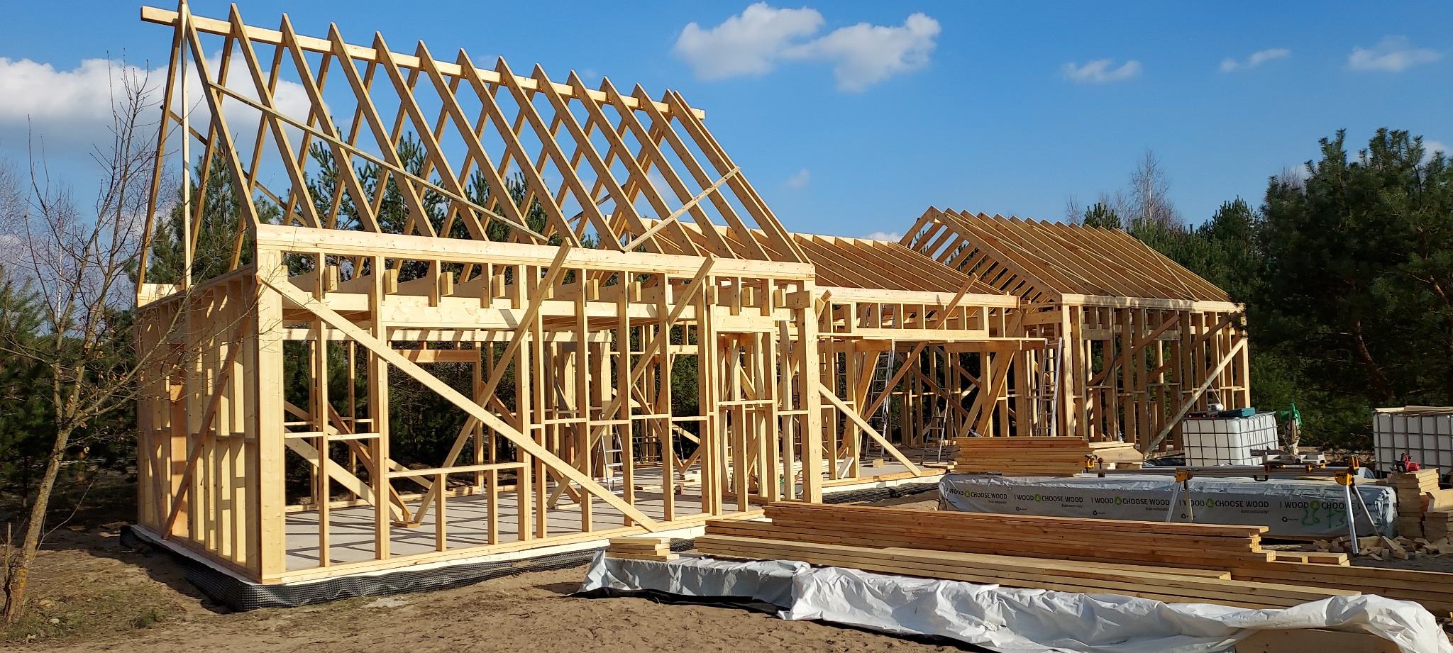 Konstrukcja szkieletowa domu w trakcie budowy, widoczne drewniane elementy ścian i dachu na tle lasu i błękitnego nieba z chmurami, z materiałami budowlanymi na pierwszym planie.