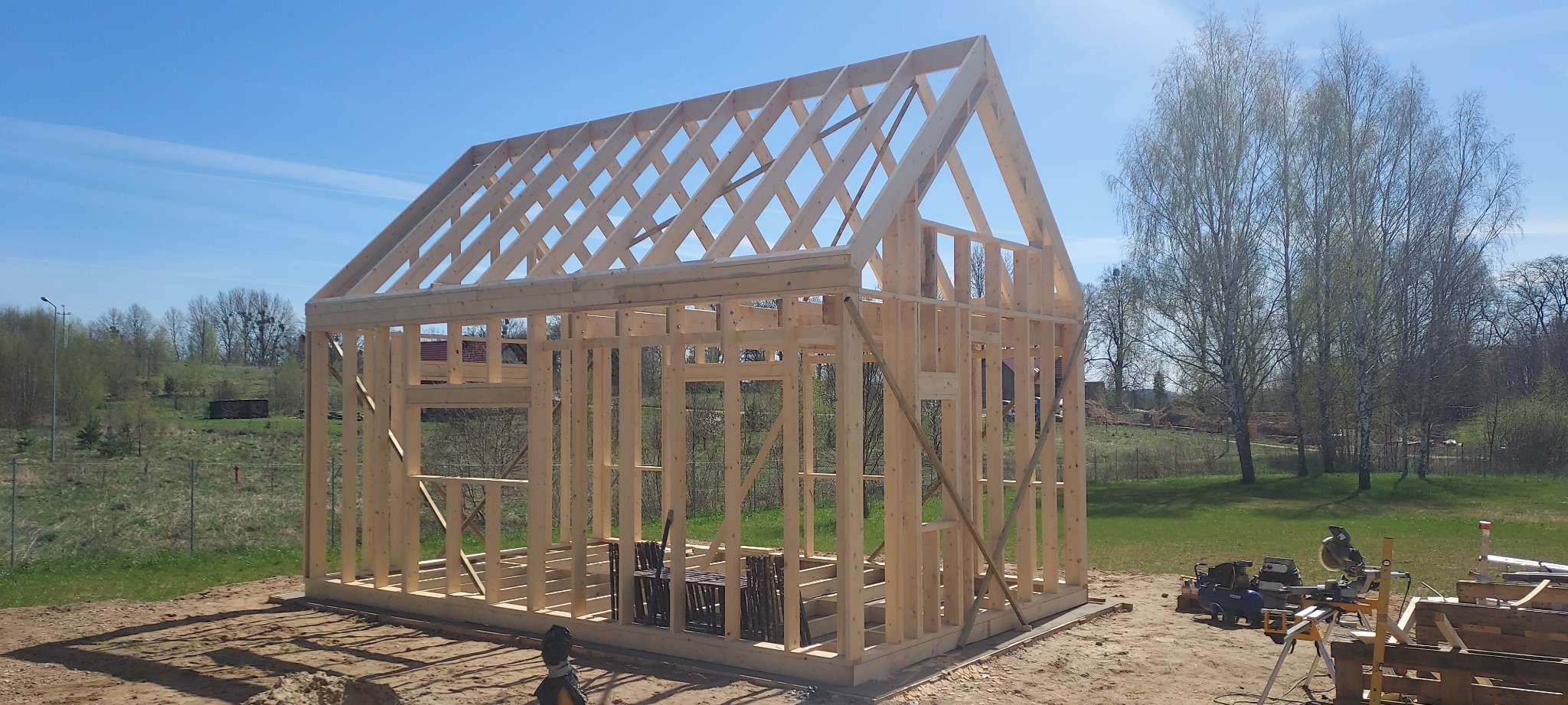 Wznoszenie drewnianej konstrukcji szkieletowej domu na fundamencie, widoczne narzędzia budowlane i otaczający krajobraz.