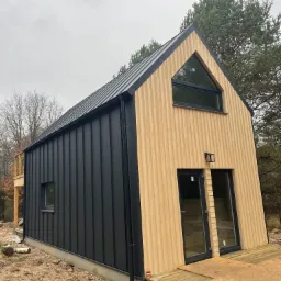 Nowoczesny dom z czarną blachą trapezową i jasną elewacją drewnianą, z asymetrycznym oknem trójkątnym na szczycie, otoczony drzewami na działce w trakcie prac wykończeniowych.