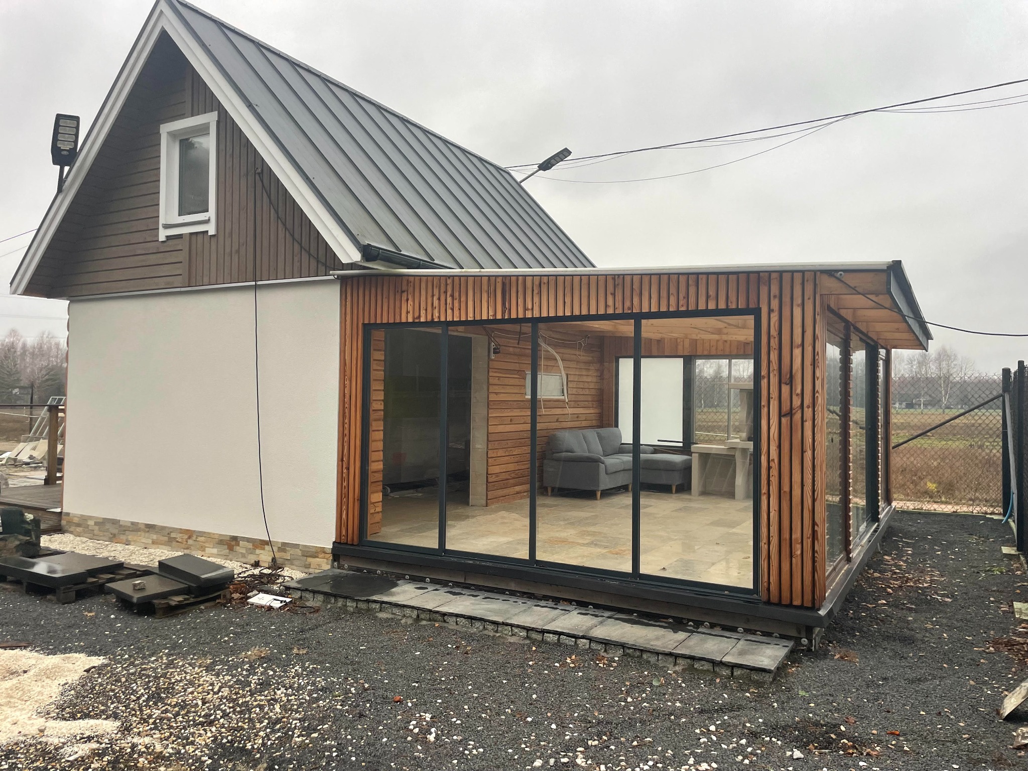 Nowoczesny dom z przeszkloną dobudówką z drewnianą elewacją, widok na szarą sofę i stolik wewnątrz.