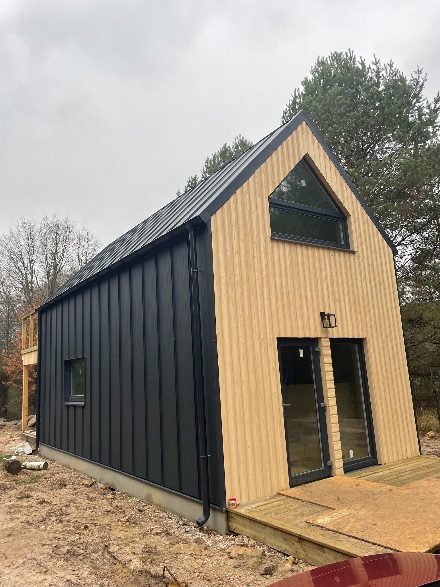 Nowoczesny dom z czarną blachą trapezową i jasną elewacją drewnianą, z asymetrycznym oknem trójkątnym na szczycie, otoczony drzewami na działce w trakcie prac wykończeniowych.