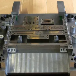 Metalowa forma do wykrawania z licznymi otworami o różnych kształtach, widoczne sprężyny i śruby mocujące, całość umieszczona na kartonowym podłożu.