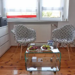 Jasne biuro z dwoma krzesłami w geometryczny wzór, szklanym stolikiem z owocami, biurkiem z drukarką i regałem na dokumenty, tworzące potencjalną przestrzeń do spotkań z klientami.