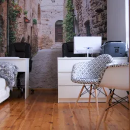 Biuro z fototapetą przedstawiającą włoską uliczkę, białe biurka, krzesła w stylu skandynawskim i lakierowana drewniana podłoga.