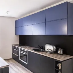 Nowoczesna kuchnia z granatowymi szafkami wiszącymi, czarnymi szafkami dolnymi i blatem imitującym drewno. Widoczne dwa piekarniki w zabudowie, zlew, suszarka do naczyń i czajnik.