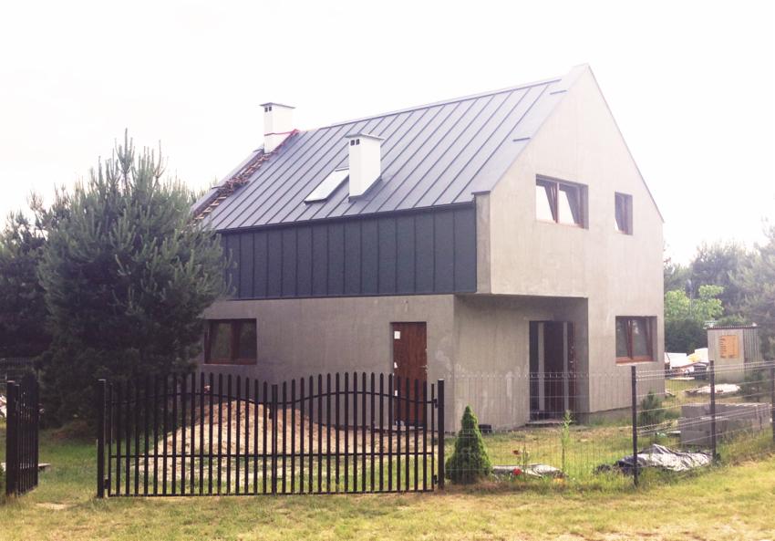 Dwukondygnacyjny dom w stanie surowym zamkniętym z ciemnoszarą blachą na dachu, czarnym panelem elewacyjnym i czarnym metalowym ogrodzeniem frontowym, widoczne drzewa i trawa.