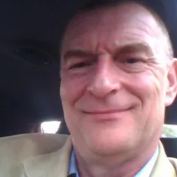 Portret uśmiechniętego mężczyzny w beżowej marynarce i błękitnej koszuli, z widoczną częścią wnętrza samochodu w tle, wykonany w naturalnym oświetleniu.